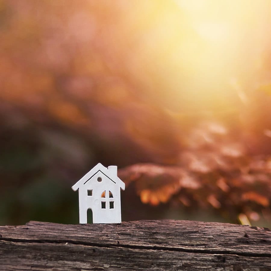 A small white house figurine sits on a wooden surface with a blurred, sunlit outdoor background.